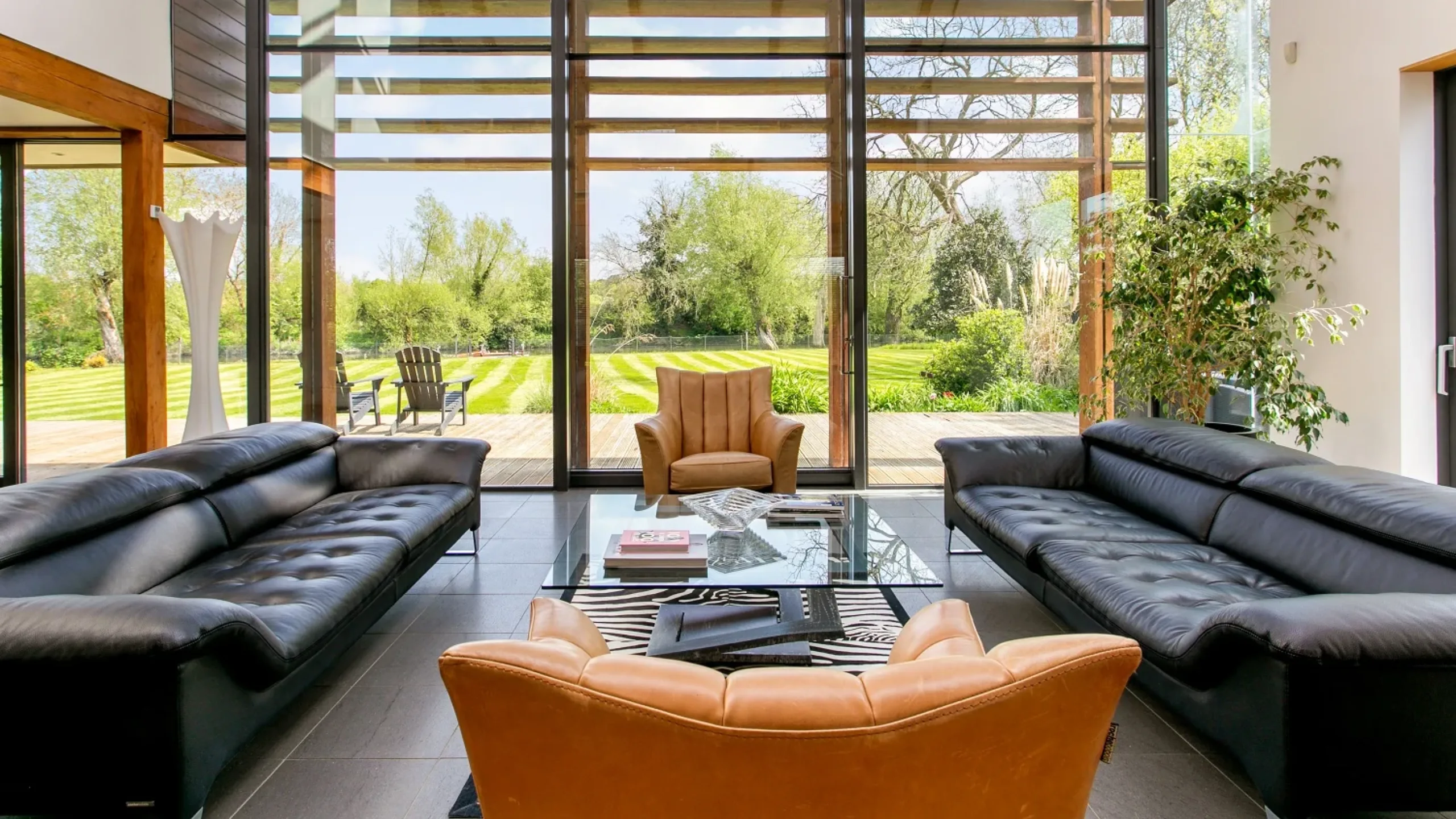 Living room with black leather sofas, orange armchairs and a clear coffee table with large sliding doors leading out to a garden