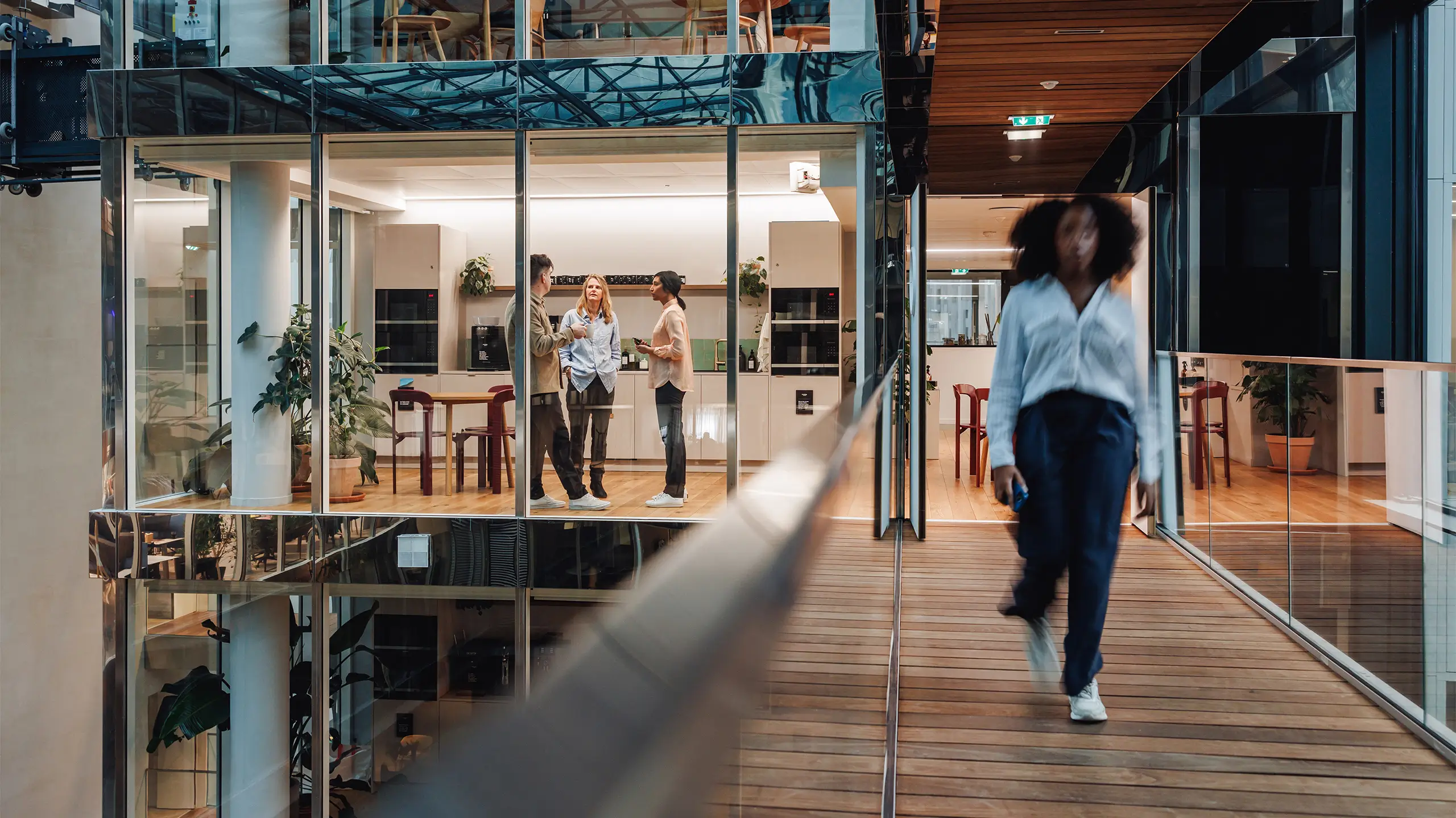 Male and female business professionals talk animatedly in a sleek, glass-walled office cafe, reflecting diversity in a corporate setting. Another walks by, blur indicative of a dynamic environment.